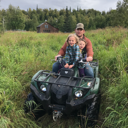 Two girls and their dad riding an ATV through a grassy area
