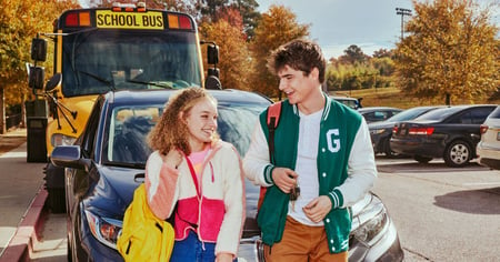 Teenage boy and girl talking while leaning on car in front of school bus.