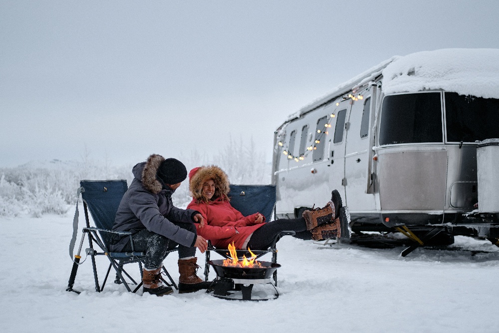 Couple sitting by fire in the winter with their 5th wheel in the background